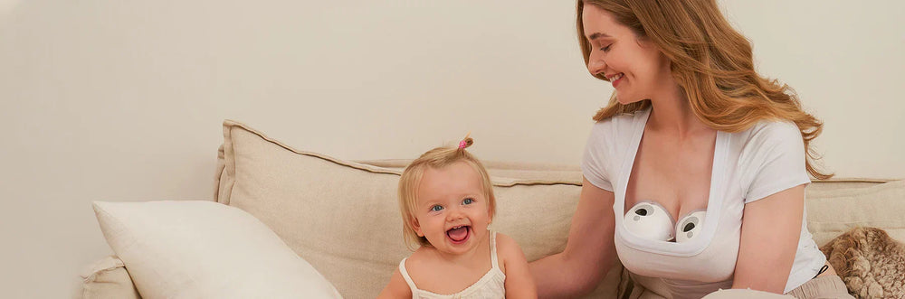 A mother wearing a wearable breast pump is sitting on a couch, bonding with her baby.