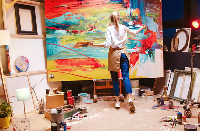 Back view of a woman artist in an apron working on a vibrant abstract painting, surrounded by art supplies in a creative studio setting.