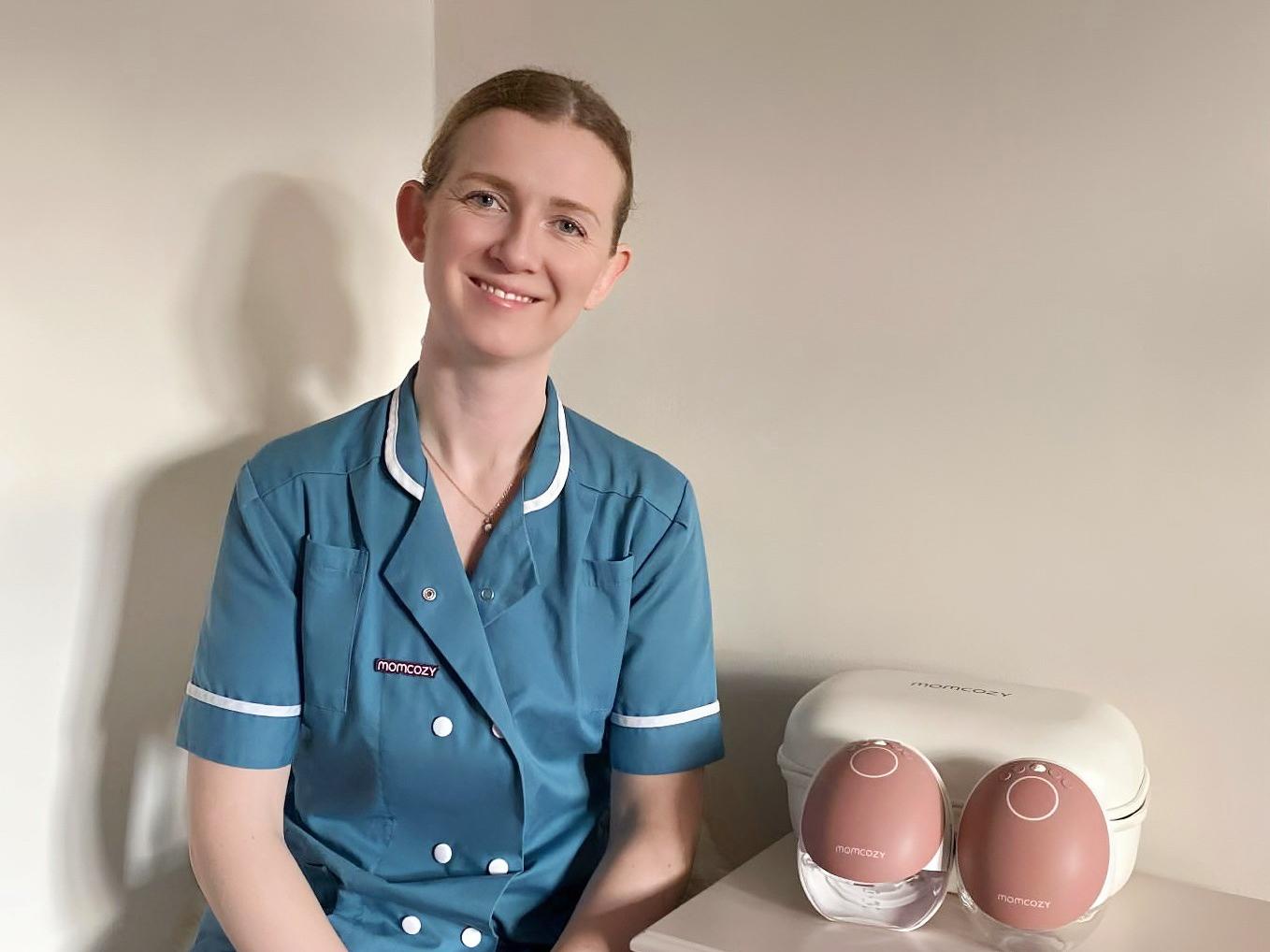 Smiling healthcare worker in teal medical scrubs with Momcozy branding, seated next to white nightstand featuring two rose gold Momcozy wearable breast pumps in their storage case