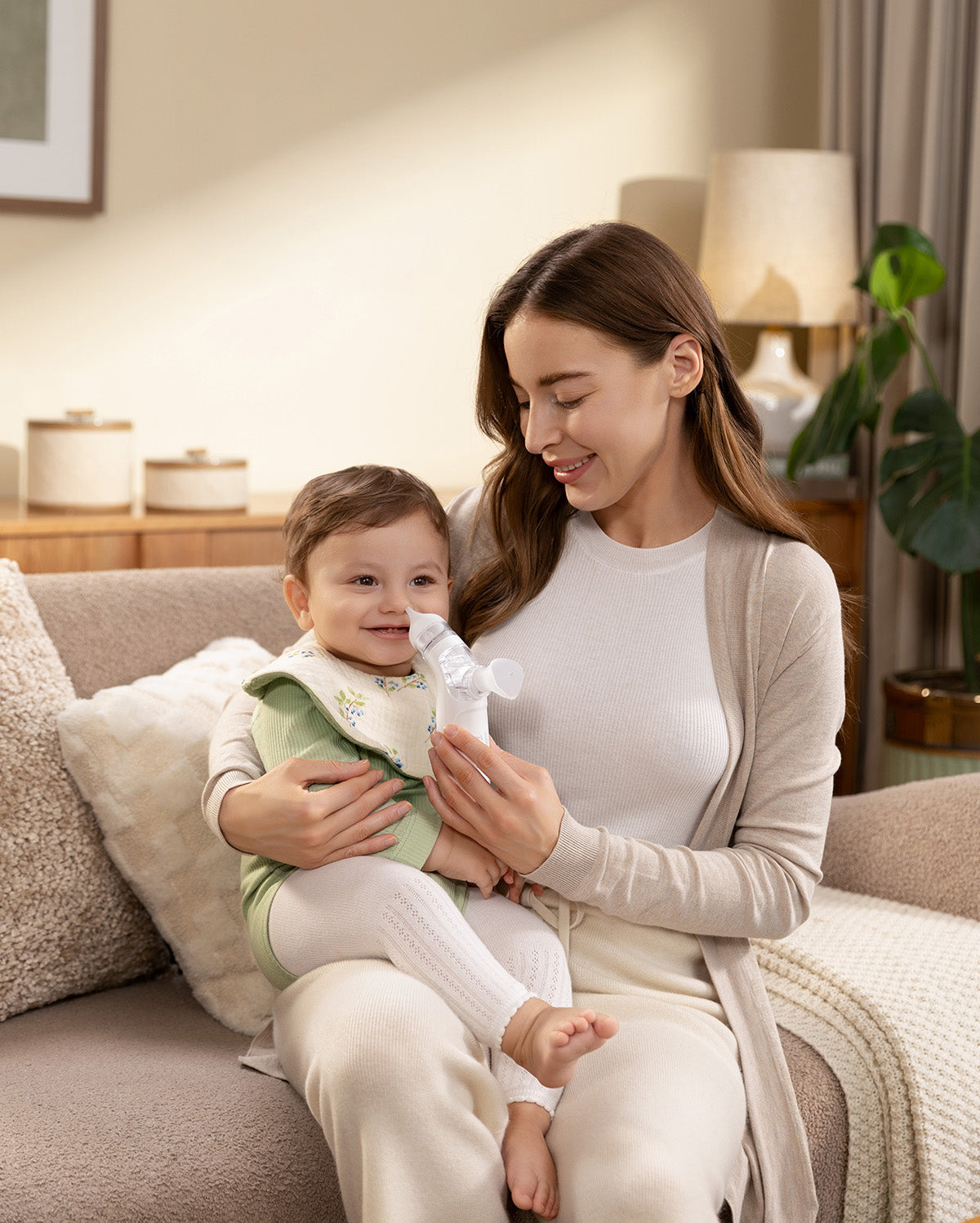 Momcozy BreezyClear electric nasal aspirator being used on a baby while sitting on a couch.